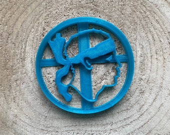 May include: A blue plastic cookie cutter shaped like a person's head with a headband. The cookie cutter is round and has a cross-shaped design in the center.