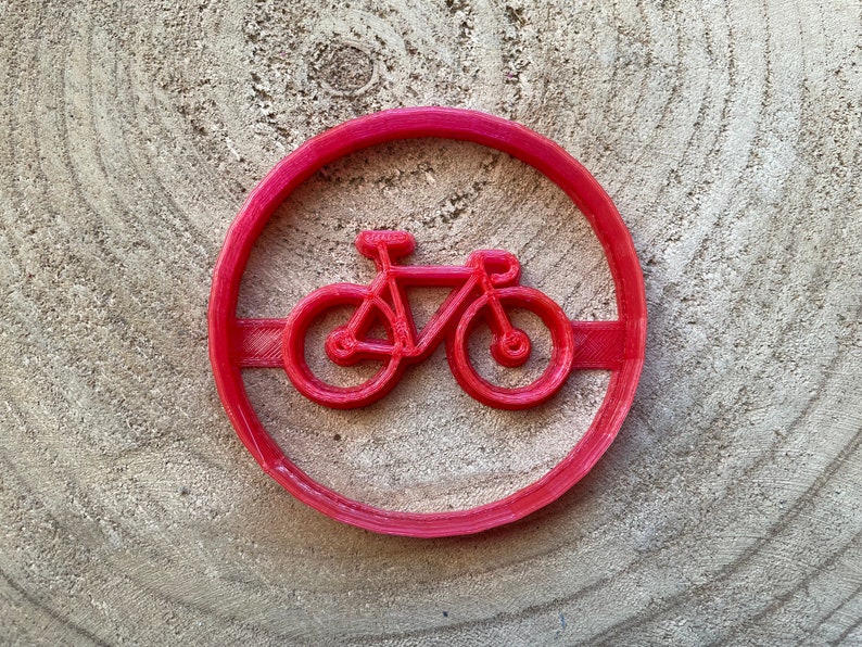 May include: Red plastic cookie cutter shaped like a bicycle. The bicycle has a simple design with two wheels, handlebars, and a seat.