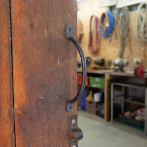 May include: Close-up of a wooden door with a black metal handle and latch. The door is made of weathered, reddish-brown wood. The handle is a curved bar, and the latch is a small, decorative piece. The background shows a workshop.
