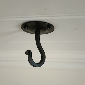 May include: Black metal ceiling hook with a decorative curved design. The hook is attached to a round metal plate.