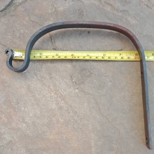 May include: Hand-forged metal plant hanger, approximately 25 cm long. The hanger has a curved top with a decorative loop and a straight vertical section with two holes for mounting. The metal has a dark, rustic finish.