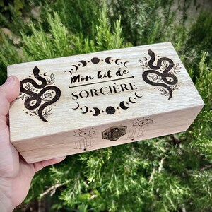 Puede incluir: Una caja de madera de color claro con un cierre metálico, con diseños grabados de serpientes, lunas y estrellas. El texto "Mon kit de SORCIÈRE" está centrado en la tapa. La caja se sostiene en una mano sobre un fondo de follaje verde.