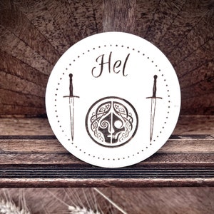 Peut inclure: Une pancarte ronde en bois blanc avec le mot "Hel" et une image stylisée d'un visage de femme avec des épées de chaque côté.
