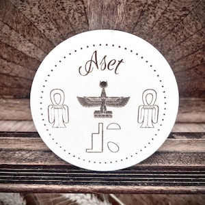 Puede incluir: Un posavasos circular blanco con una imagen en blanco y negro de la diosa egipcia, Aset, con el texto "Aset" en cursiva.