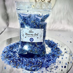 Op de afbeelding: Een doorzichtige zak gevuld met blauw badzout, met het label "Sel Magique Bleu Mauvais Oeil". De zak staat op een spiegelend oppervlak, omringd door verspreid blauw zout. Een ronde sticker met een hemels ontwerp is op de achtergrond.