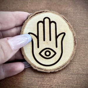 Può includere: Un sottobicchiere di legno con un design a mano di hamsa in bianco e nero con un occhio al centro.