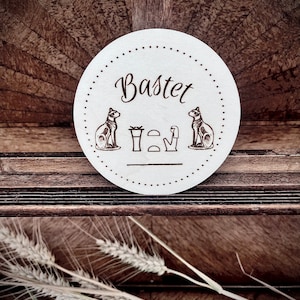 Puede incluir: Un posavasos redondo blanco con una imagen en blanco y negro de la diosa egipcia Bastet. El posavasos tiene un borde negro punteado y el texto "Bastet" en cursiva.