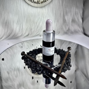 Peut inclure: Une bouteille noire et argentée avec un compte-gouttes violet clair, posée sur une surface réfléchissante. La bouteille est entourée de pierres noires et de clous rouillés croisés. Un emblème circulaire noir et blanc est en arrière-plan.