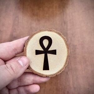 Puede incluir: Un posavasos de madera con un símbolo Ankh negro, un jeroglífico egipcio tradicional, en el centro.