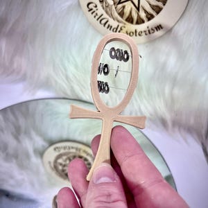 Puede incluir: Un colgante beige en forma de ankh con anillos negros suspendidos de alambres. El colgante se sostiene en una mano, con un fondo borroso que presenta un diseño circular con el texto "Girl and Esoterism".