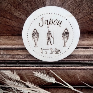 Puede incluir: Un posavasos redondo de madera con fondo blanco y tinta negra. El posavasos presenta un dios egipcio con un bastón y dos figuras con colas de pez. El texto "Inpou" está escrito en cursiva en la parte superior del posavasos.