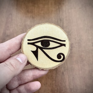 Puede incluir: Un posavasos de madera con una imagen en blanco y negro del Ojo de Horus, un símbolo egipcio antiguo.