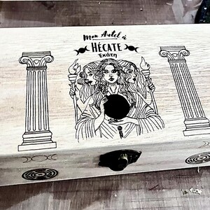 Op de afbeelding: Een lichtgekleurde houten kist met een scharnierend deksel, met een gegraveerd ontwerp. Het ontwerp omvat drie vrouwelijke figuren, kolommen en de tekst "Mon Autel à Hécate". Een zwarte sluiting zekert de kist.