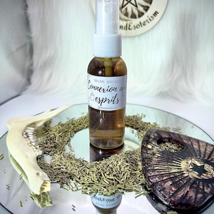 Puede incluir: Un frasco rociador de vidrio marrón con una tapa blanca, etiquetado como "Brume Magique Connexion aux Esprits", se encuentra sobre una superficie espejada. Hierbas secas, un hueso y una pieza decorativa de madera rodean el frasco. Un emblema circular está en el fondo.