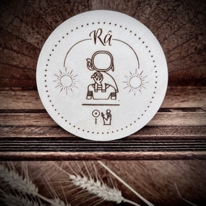 Puede incluir: Un posavasos redondo de madera blanco con la palabra "Rá" y una imagen estilizada del dios egipcio Ra, con un sol a cada lado de la imagen.