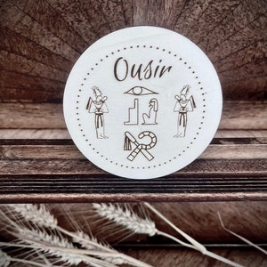 Puede incluir: Un posavasos redondo de madera con fondo blanco e ilustraciones en blanco y negro de dioses y símbolos egipcios. El texto "Ousir" está escrito en cursiva en la parte superior del posavasos.