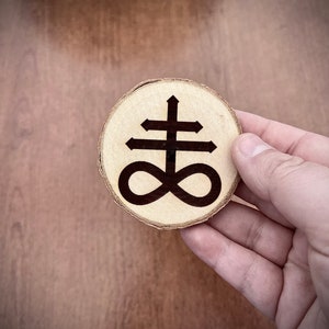 Op de afbeelding: Een houten cirkel met een zwart symbool van een kruis met een oneindigheidssymbool eronder.