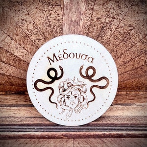 Può includere: Sottobicchiere rotondo in legno con design Medusa. La parola greca "Μέδουσα" è in alto, con un'illustrazione del volto di Medusa e dei serpenti. Il design è marrone scuro. Diametro di circa 10 cm.