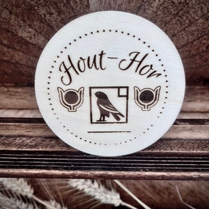 Puede incluir: Un posavasos redondo de madera clara con las palabras "Hout-Hor" en una fuente decorativa. El posavasos presenta una ilustración de un pájaro dentro de un cuadrado y dos diseños de tazas estilizadas. Un borde punteado rodea el texto y las imágenes.