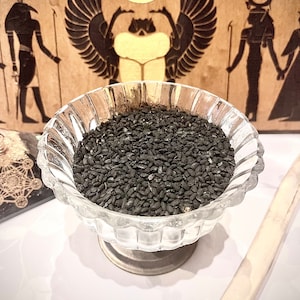 Può includere: Una ciotola di vetro piena di semi di nigella neri, noti anche come semi di cumino nero. La ciotola è su una superficie bianca. Lo sfondo presenta un pannello di legno con un design di ispirazione egizia.