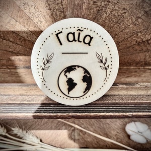 Può includere: Un cartello rotondo in legno con la parola "Gaia" scritta in lettere nere. Il cartello presenta anche una silhouette nera della Terra e due piccoli rami con foglie su entrambi i lati della parola.
