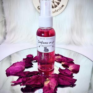 Puede incluir: Un frasco rociador rosa con tapa blanca, etiquetado "Brume Magique Confiance en soi" con un gráfico. Pétalos de rosa secos rodean el frasco sobre una superficie reflectante. Se ve un fondo blanco con un diseño circular.