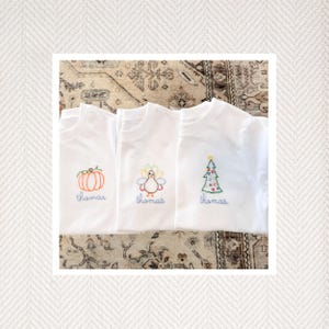 May include: Three white t-shirts with embroidered designs. One shirt features an orange pumpkin, another a turkey, and the last a Christmas tree. Each shirt has the name "thomas" embroidered in blue thread below the design.