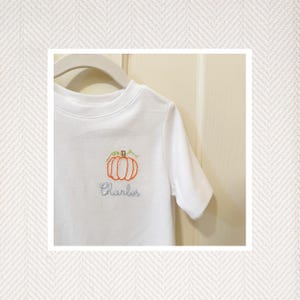 May include: White short-sleeved t-shirt with an embroidered orange pumpkin and green stem. The name "Charles" is embroidered below the pumpkin in light blue thread. The shirt is hanging on a white hanger.