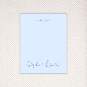 May include: A light blue notepad with the text "a note from" at the top and "Sophie Lucas" at the bottom in a dark blue script font. The notepad is rectangular and sits on a light beige patterned surface.