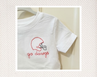 May include: A white short-sleeved t-shirt featuring red and black embroidery on the chest. The design depicts a football helmet outlined in red with black face mask details, accompanied by the red embroidered text "go dawgs" underneath.