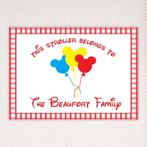May include: A white sign with a red and white checkered border. The sign says "This stroller belongs to The Beaufort Family". There are three Mickey Mouse head balloons in the center of the sign, one red, one yellow, and one blue.