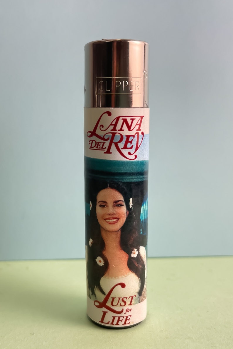 May include: A silver Clipper lighter with a white label featuring a photo of Lana Del Rey and the text "Lana Del Rey" and "Lust for Life".
