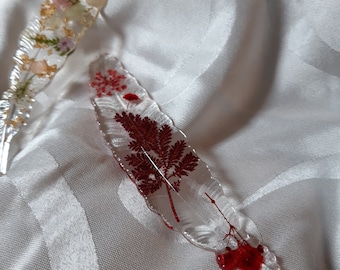 May include: Two clear resin bookmarks with pressed flowers and leaves. One bookmark features gold leaf and pink and white flowers. The other bookmark features red flowers and leaves.