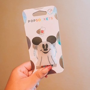 May include: A white and black PopSockets phone grip featuring a ghost Mickey Mouse design with glitter accents. The packaging says "PopSockets" and "Pull twice to grip".