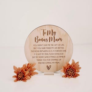 May include: A wooden plaque with the text "To My Bonus Mum" and a heartfelt message about the bond between a stepparent and child. The plaque is on a small wooden stand and surrounded by orange flowers.
