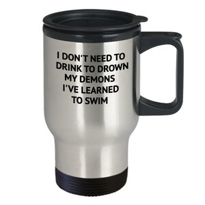 May include: Stainless steel travel mug with a black lid and handle. The mug has a black inscription that reads "I don't need to drink to drown my demons I've learned to swim."