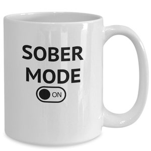 Może przedstawiać: Biały ceramiczny kubek z napisem "SOBER MODE" w czarnym kolorze i grafiką włącznika "ON". Kubek ma zakrzywiony uchwyt i jest przeznaczony do napojów.