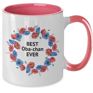 Puede incluir: Taza de café de cerámica blanca con borde y asa de color rosa. La taza presenta una corona de flores con flores rojas y azules y el texto "BEST Oba-chan EVER".