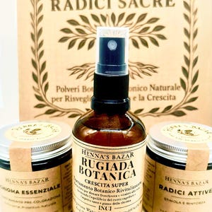 Op de afbeelding: Een productkit van Henna's Bazar, met een bruine spuitfles en twee potten. De spuitfles is gelabeld met "Rugiada Botanica", de potten met "Chioma Essenziale" en "Radici Attive". De achtergrond toont een bord met "Henna's Bazar" en "Radici Sacre".