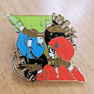 May include: A gold-coloured enamel pin featuring two cartoon characters. The character on the left is wearing a blue hoodie and headphones, while the character on the right is wearing a red hoodie and green hair. Both characters are pointing at each other with their index fingers. The background is a cityscape with gold accents.