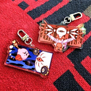 May include: Two colorful acrylic keychains featuring anime characters. One keychain is red, white, and black with a character with a single eye and wings. The other keychain is blue, black, and pink with a character with a pink body and a single eye.