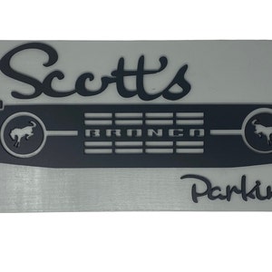May include: A silver metal sign with black lettering that reads "Scott's Parking" with a black silhouette of a Ford Bronco grille in the center.