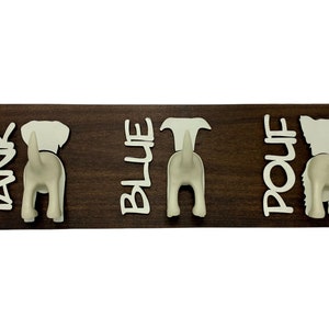 K&ouml;nnte beinhalten: Ein Wand-Haken aus Holz mit drei wei&szlig;en Haken in Form von Hundepopos. Die Haken sind mit "Tank", "Blue" und "Pouf" beschriftet.