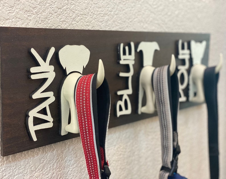 K&ouml;nnte beinhalten: Ein braunes, h&ouml;lzernes Hunde-Leinenregal mit drei wei&szlig;en, hundeartigen Haken. Die Haken sind mit "Tank", "Blue" und "Hope" beschriftet. Drei Leinen h&auml;ngen an den Haken.