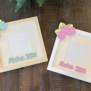 Puede incluir: Dos marcos de fotos cuadrados, uno de madera natural y otro blanco, cada uno adornado con detalles florales y el texto "Aloha 2024". Los marcos tienen un paspartú color crema y están sobre un fondo de madera.