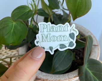 May include: A white sticker with the words "Plant Mom" in a green, decorative font. The sticker is surrounded by green leaf illustrations. The sticker is held in front of a potted plant with green leaves.