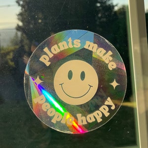 May include: A holographic sticker with a rainbow iridescent background and a white smiley face. The sticker says "Plants make people happy" in a retro font.