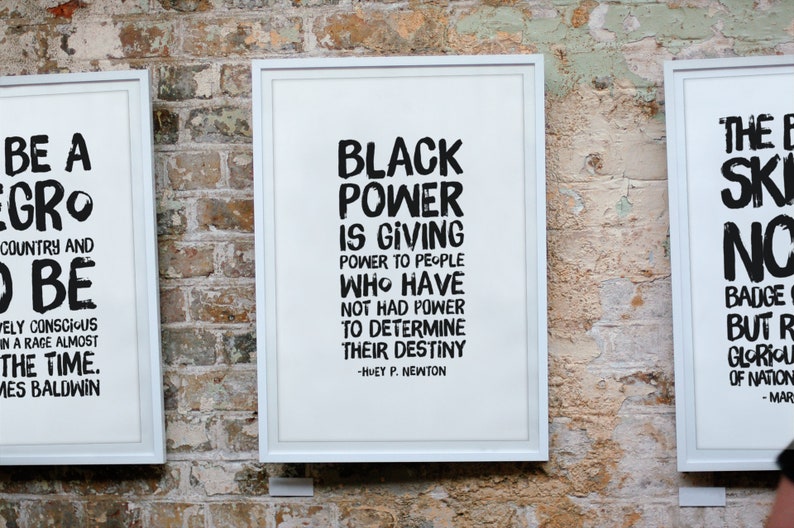 May include: Three framed prints with black text on a white background. The first print says "Be a Negro, Country and To Be" by James Baldwin. The second print says "Black Power is giving power to people who have not had power to determine their destiny" by Huey P. Newton. The third print says "The Black Skin, No Mask" by Frantz Fanon.