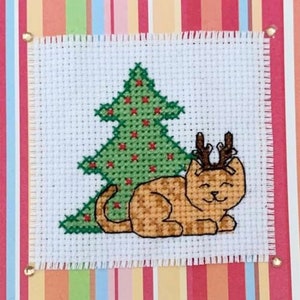 May include: A cross-stitch design featuring a brown cat wearing reindeer antlers, sitting next to a green Christmas tree with red ornaments.