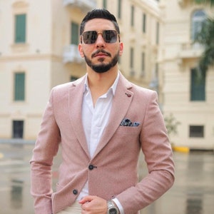 Puede incluir: Chaqueta rosa claro con cierre de dos botones, combinada con una camisa blanca y pantalones color crema. La chaqueta tiene un pañuelo de bolsillo y se usa con un reloj de pulsera plateado y gafas de sol. El fondo es una escena urbana borrosa.